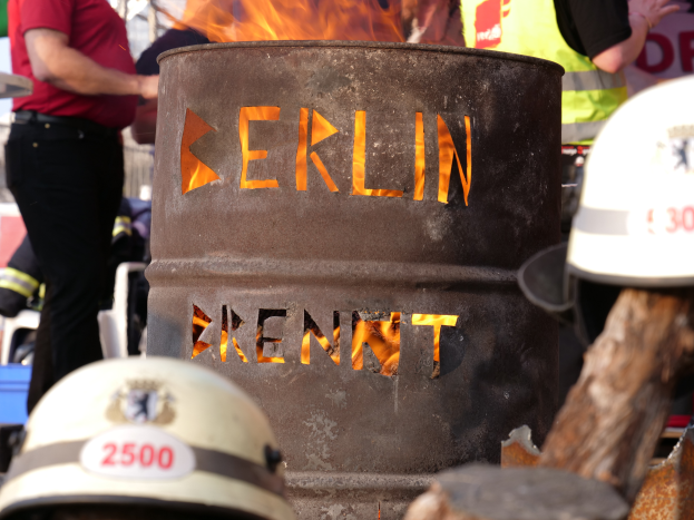 Eine Gruppe von Menschen um ein Feuerhydranten-Schild herum, auf dem 'Berlin' steht, mit einem brennenden Fass im Vordergrund, Helme auf dem Boden und ein hölzernes Objekt auf der rechten Seite.