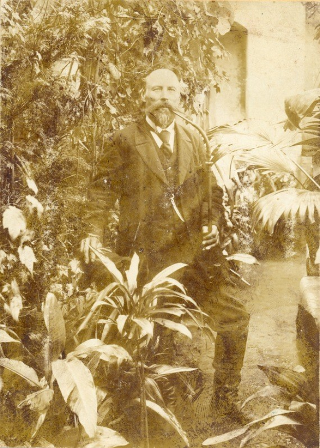 Altes Schwarz-Weiß-Foto eines Mannes in Anzug und Krawatte, der in einem Garten mit Pflanzen und Bäumen im Hintergrund und einer Wand im Hintergrund steht.
