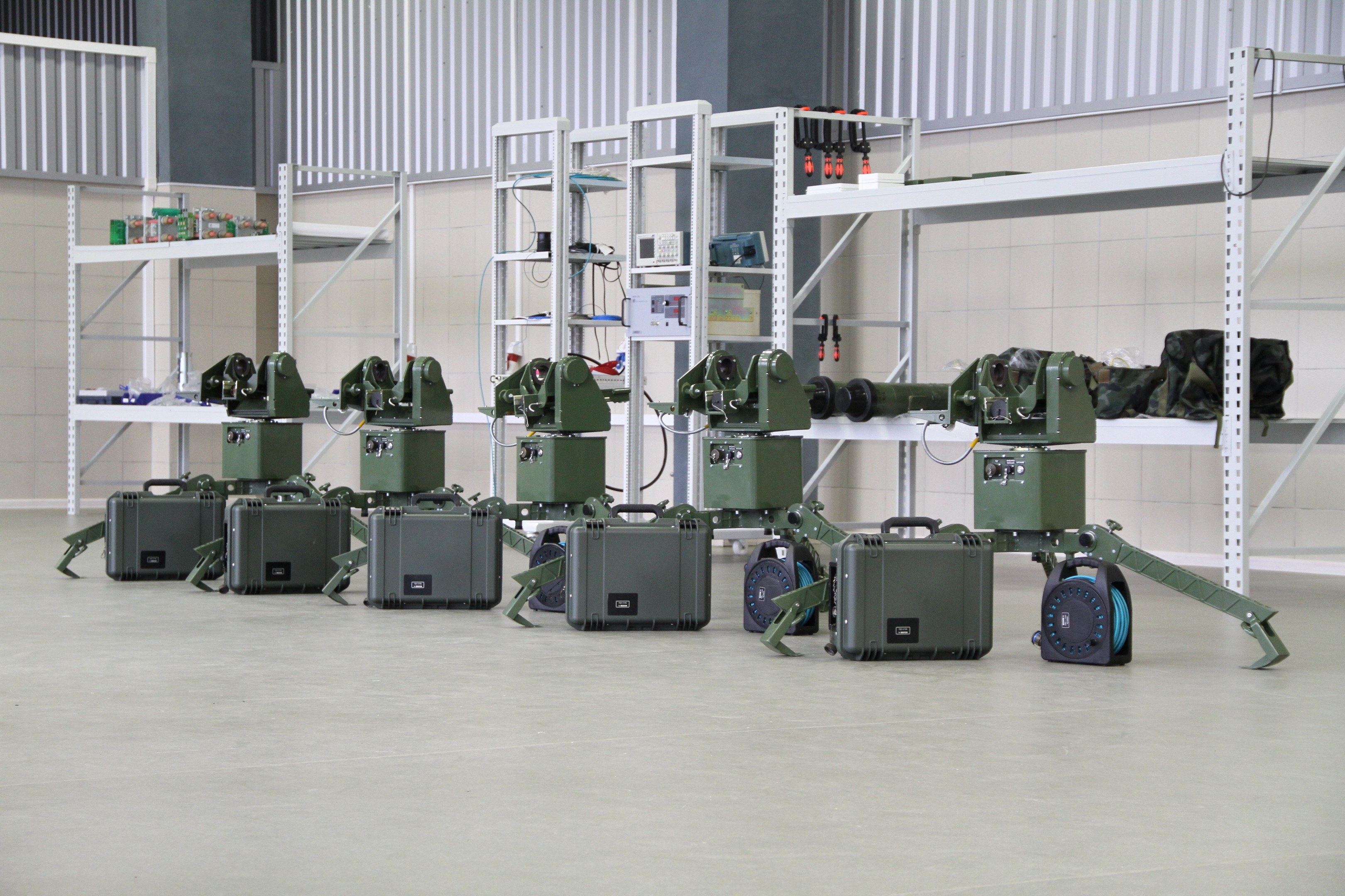 Eine Gruppe von Milit├Ąrrobotern auf einem Boden mit Koffern und anderen Gegenst├Ąnden auf umliegenden Gestellen, vor einer Wand mit einem Gel├Ąnder im Hintergrund.