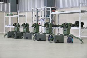 Eine Gruppe von Milit├Ąrrobotern auf einem Boden mit Koffern und anderen Gegenst├Ąnden auf umliegenden Gestellen, vor einer Wand mit einem Gel├Ąnder im Hintergrund.