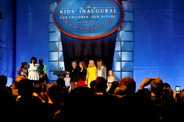 Eine Gruppe von Menschen auf einer Bühne, einige halten Mobiltelefone, mit einem Vorhang und einem Textbrett im Hintergrund bei einer Kindereröffnungsveranstaltung.