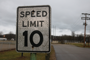 Ein Tempolimit-Schild am Straßenrand, umgeben von Gras, Strommasten mit Drähten, einem Zaun, Häusern, Bäumen und einem bewölkten Himmel im Hintergrund.