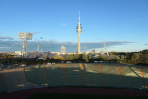 Olympiastadion in Berlin, Deutschland, mit dem Fernsehturm (Fernsehturm) im Hintergrund, umgeben von Bäumen, Gebäuden und Lichtern unter einem bewölktem Himmel.