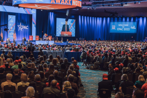 Eine große Menschenmenge sitzt auf Stühlen, die einer Bühne zugewandt sind, mit ein paar Menschen darauf, blaue Vorhänge und ein Bildschirm im Hintergrund und ein orangefarbenes Schild oben, das das CPAC 2020-Event am 25. Oktober ankündigt.