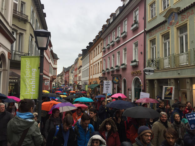 Große Gruppe von Menschen geht während einer Klimademo in Deutschland eine Straße entlang, viele halten Schirme, Taschen und Schilder, mit Gebäuden und einem Lichtmast im Hintergrund.