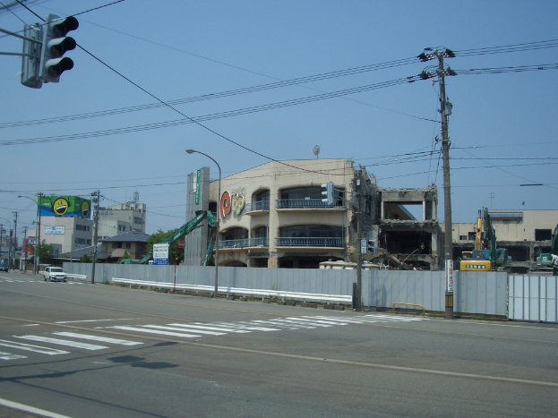 Eine Stadtstraße mit Fahrzeugen, Strommasten, Laternenmasten und einer Ampel auf der linken Seite, Gebäuden, Bäumen und dem Himmel im Hintergrund und einem Gebäude in derForeground, das abgerissen wird.