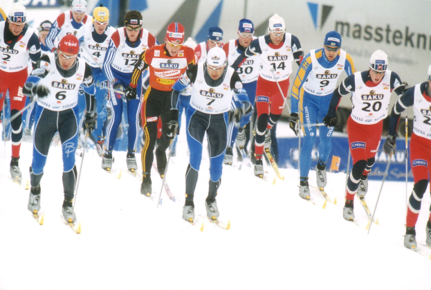 Gruppe von Menschen, die eine schneebedeckte Abh"angigkeit hinunterfahren, tragen Skihelm, M"utze, Handschuhe und halten Ski-Stöcke mit Rennstart-Tafeln im Hintergrund.