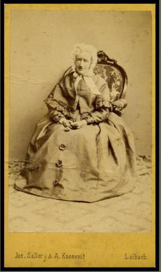 Ein altes Foto einer Frau mit sanftem Gesichtsausdruck, die ein helles Kleid und einen gemusterten Schal trägt und auf einem Stuhl sitzt, mit ihren Händen auf dem Schoß ruhend und ihrem Haar zu einem ordentlichen Dutt gebunden, in einem Foto Rahmen präsentiert.