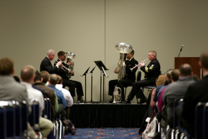 Eine Gruppe von Männern, die auf einer Bühne Blasinstrumente spielen, mit Notenständern, einem Podium mit Mikrofon und einem Publikum, das auf Stühlen sitzt, sowie einer Wand im Hintergrund.