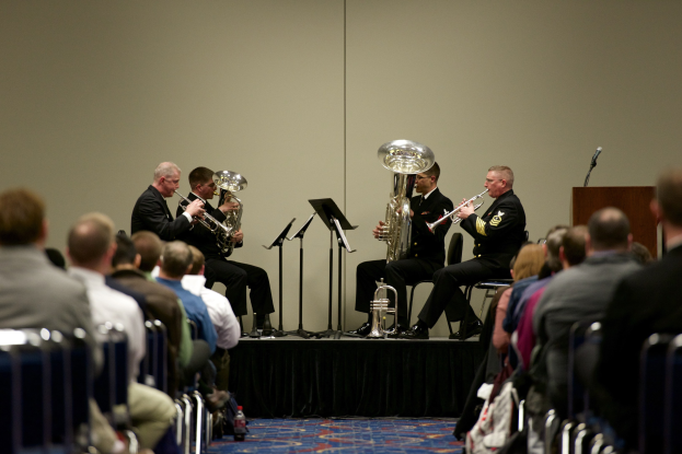 Eine Gruppe von Männern, die auf einer Bühne Blasinstrumente spielen, mit Notenständern, einem Podium mit Mikrofon und einem Publikum, das auf Stühlen sitzt, sowie einer Wand im Hintergrund.