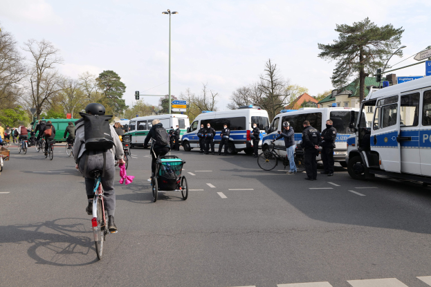 Eine Gruppe von Menschen, die auf Fahrrädern eine Straße entlangfahren, mit Polizeiwagen, anderen Fahrzeugen, Bäumen, Straßenschildern, Gebäuden und einem bewölktem Himmel.