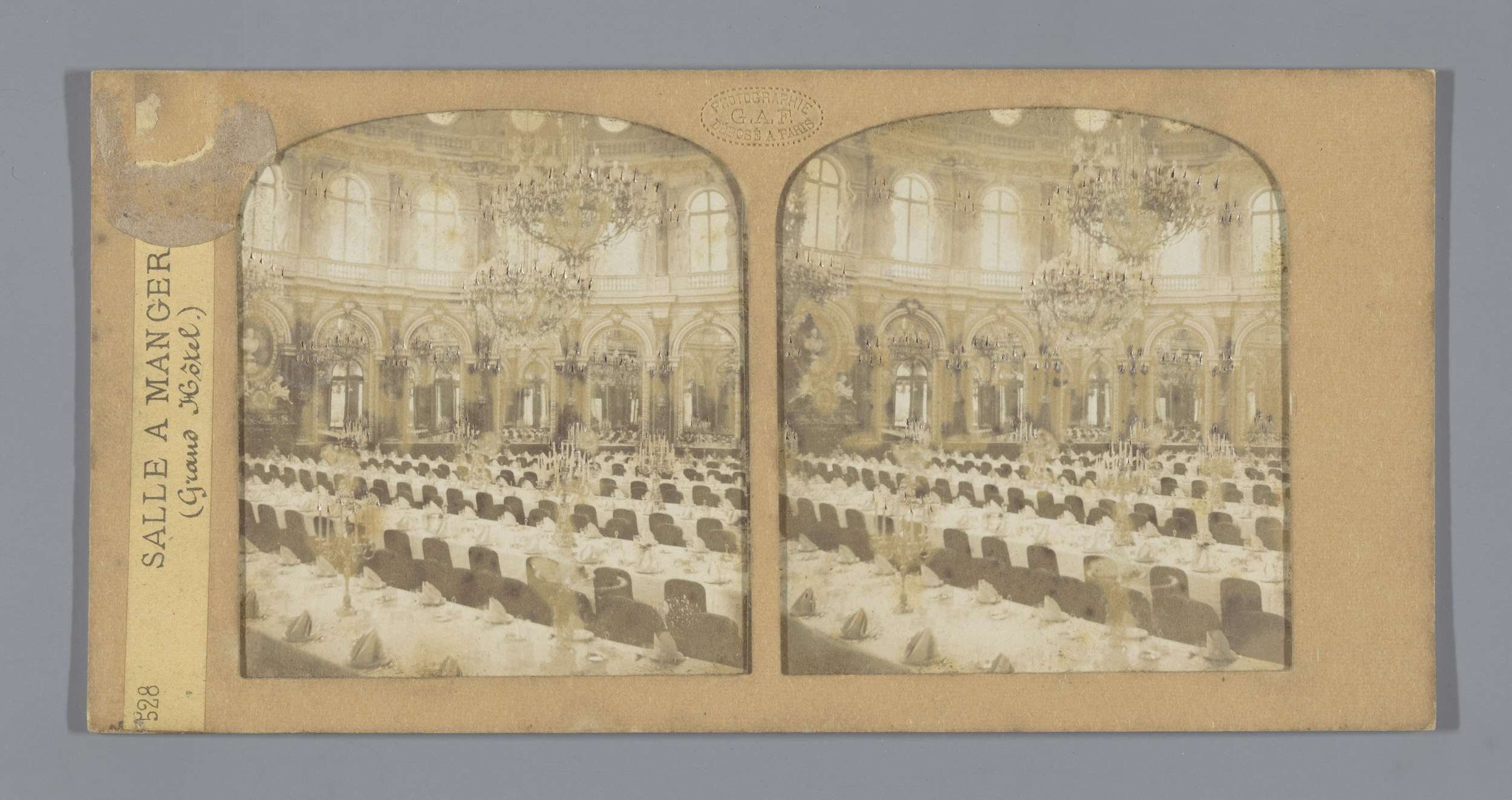 Ein altes Foto eines prächtigen Hotel-Speisesaals mit Tischen, Stühlen, Kronleuchtern und Fenstern, das den Text "Sale a Manger, Grand Hotel" auf der linken Seite zeigt.