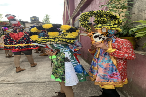 Eine Gruppe von Menschen in bunten Kostümen steht zusammen vor einem Gebäude, mit verschiedenen Gegenständen, Bäumen, Gebäuden und einem klaren blauen Himmel im Hintergrund.