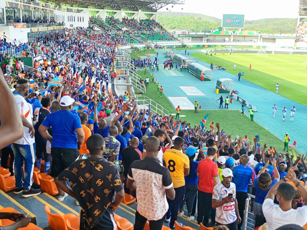 Eine große Menschenmenge versammelt sich vor einem eingezäunten Fußballfeld, wobei einige auf Stühlen sitzen und andere stehen, unter einem klaren blauen Himmel mit Hügeln im Hintergrund, und warten auf ein Spiel.