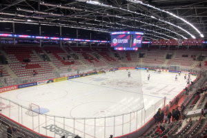 Eishockeyspiel in einer großen Arena mit Zuschauern, ein von einem Zaun umgebenes Eis, ein Display mit "St. Louis Blues vs Detroit Red Wings", Metallrahmen, Deckenbeleuchtung und Dach.