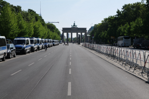 Lange Reihe von Polizeiwagen am Straßenrand vor dem Brandenburger Tor in Berlin, Deutschland, mit Fahrradfahrern und Menschen auf der Straße, Barrieren, Bäumen, einem Bogen mit Statuen im Hintergrund und sichtbarem Himmel.