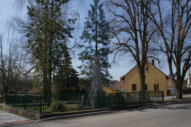 Eine Straße mit einem Holocaust-Denkmal in ihrer Mitte, umgeben von einem Metallzaun, umgeben von Gras, Bäumen, Häusern, einem Straßenpfahl und einem bewölkten Himmel.