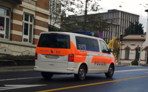 Ein Polizeiwagen fährt eine Straße mit hohen Gebäuden, Bäumen, Pflanzen, Pfählen, Verkehrszeichen und Schildern entlang, mit dem Himmel im Hintergrund.