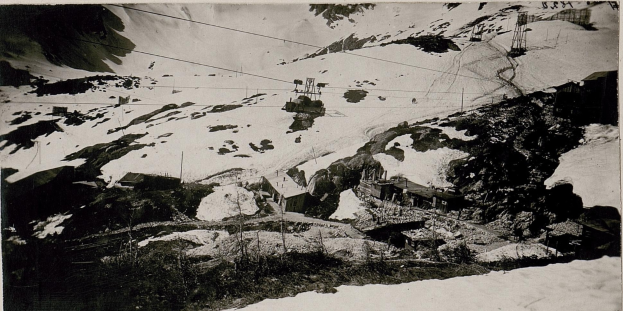 Ein Schwarz-Weiß-Foto eines Skilifts, der einen schneebedeckten Berg hinauffährt, mit Häusern, Pfählen und Drähten im Vordergrund und Text unten.