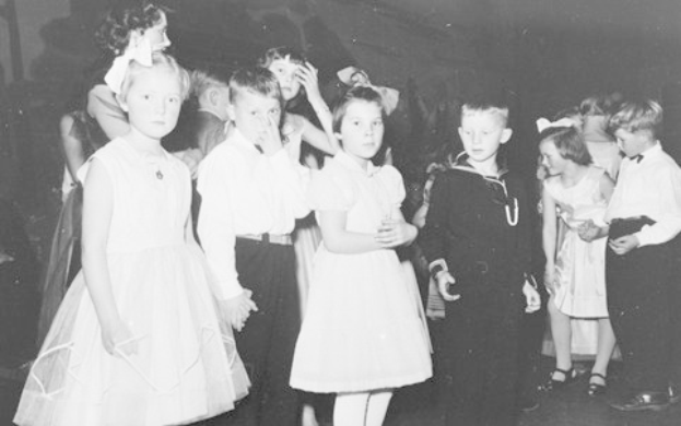 Schwarzes und weißes Bild einer Gruppe von Kindern, die auf einer Tanzfläche stehen, lächeln und fröhlich aussehen, mit einer Wand im Hintergrund.