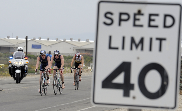 Eine Gruppe von Radfahrern mit Helmen fährt auf einer Straße mit einem Geschwindigkeitsbegrenzungsschild, Gebäuden und Bäumen im Hintergrund unter einem klaren blauen Himmel.