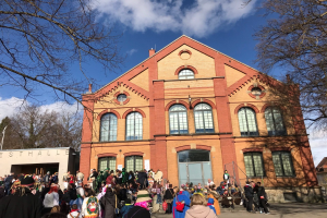Eine große Gruppe von Menschen steht vor einem mehrstöckigen Gebäude mit Fenstern und Türen, umgeben von Bäumen unter einem bewölkten Himmel, einige tragen Mützen und tragen Taschen.