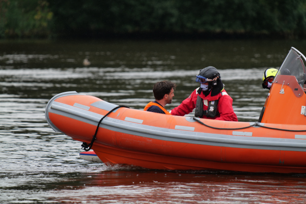 Zwei Personen in einem orangen Rettungsboot auf dem Wasser, mit Schwimmwesten und Helmen, mit Bäumen im Hintergrund.