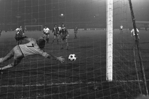 Ein Schwarz-Weiß-Foto eines Fußballspielers, der vor einem Tor einen Ball hält, umgeben von einer Gruppe von Menschen, die auf dem Boden stehen, das Torpfosten ist auf der rechten Seite des Fotos zu sehen, sowie Werbetafeln und Lichter im Hintergrund.