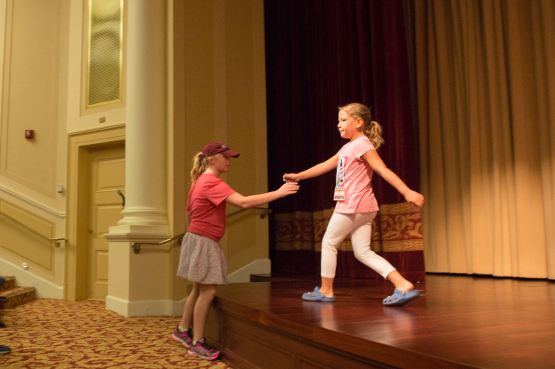 Zwei junge Mädchen tanzen auf einer Bühne vor einem Vorhang, mit einer Person, die links von ihnen auf einem Stuhl sitzt, einer Säule und einer Wand im Hintergrund.