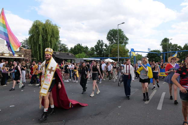 Eine Gruppe von Menschen marschiert bei der Pride Parade 2018, einige halten Musikinstrumente und tragen Mützen, und tragen eine Regenbogenfahne eine Straße mit Laternenpfählen, Bäumen, Hütten und einem bewölkten Himmel im Hintergrund.