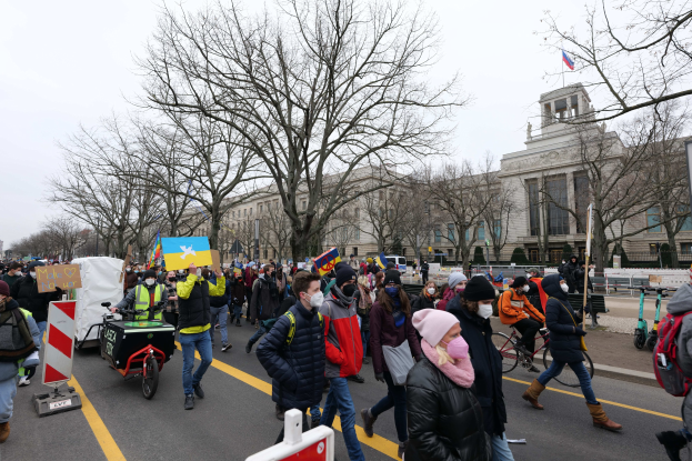 Eine große Gruppe von Menschen marschiert auf einer Straße in Washington, D.C. und hält Schilder und Fahnen, einige fahren Fahrräder, mit Bäumen, Schildern und einem klaren blauen Himmel im Hintergrund.