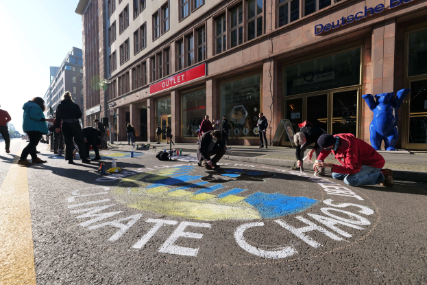 Menschen sitzen vor einem Gebäude mit Fenstern und Namensschildern während einer Klimawandel-Demo in Berlin, umgeben von Flaschen und anderen Gegenständen, mit Bäumen und einem klaren blauen Himmel im Hintergrund.