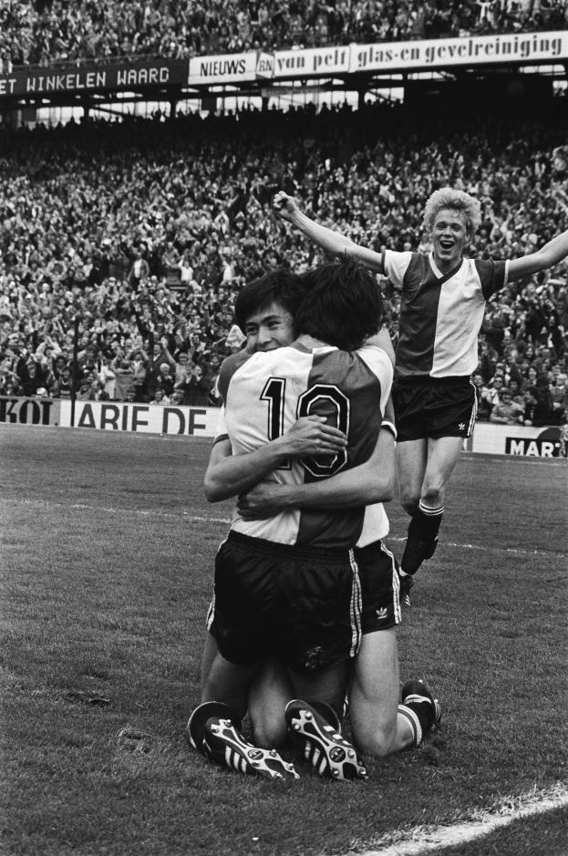 Drei Fußballspieler feiern auf dem Boden nach einem Tor, mit jubelnder Menge und schwenkenden Fahnen in einem schwarz-weißen Stadion-Setting.
