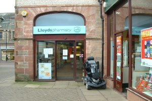 Apotheke mit einem Fahrzeug davor und einem Gebäude links daneben.