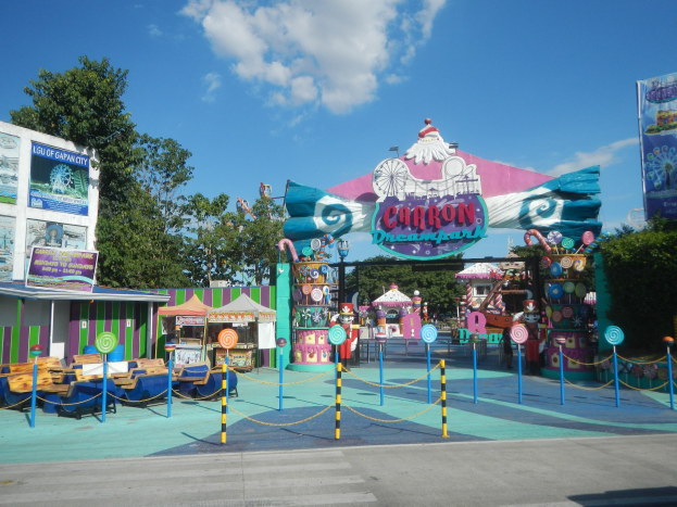 Karussell am Parkeingang mit Straße darunter, eingezäuntem Bereich, Buden, Banner, Bäumen und bewölktem Himmel im Hintergrund.
