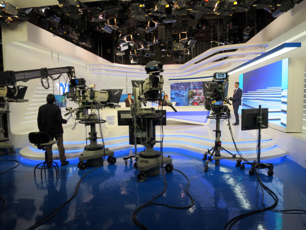 Ein TV-Studio mit mehreren Kameras auf dem Boden, einer sitzenden Person im Vordergrund und einigen stehenden Individuen im Hintergrund, ausgestattet mit Deckenlampen für eine Live-Übertragung.