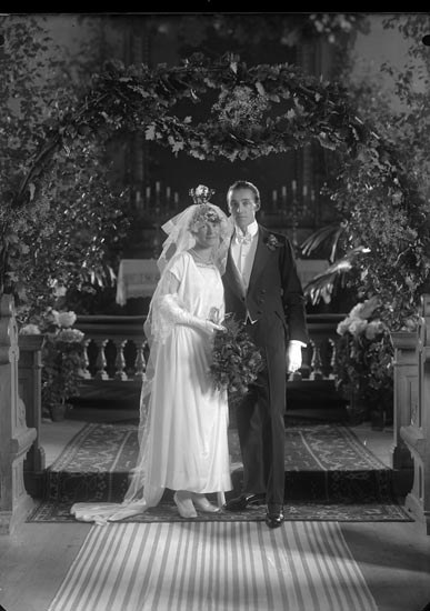 Schwarz-weiß-Foto eines Mannes und einer Frau, die unter einem Blumenschmuckbogen stehen, die Frau hält einen Strauß Blumen, mit einem Teppich auf dem Boden.