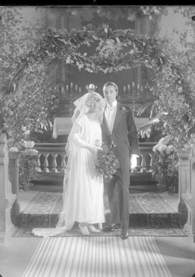 Schwarz-weiß-Foto eines Mannes und einer Frau, die unter einem Blumenschmuckbogen stehen, die Frau hält einen Strauß Blumen, mit einem Teppich auf dem Boden.