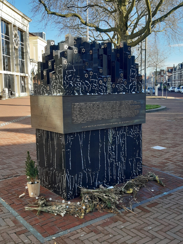 Denkmal für Holocaust-Opfer in einem Stadtplatz mit einem Baum davor, einem Topf mit einer Pflanze, verstreuten trockenen Blättern und Gebäuden, Fahrzeugen, Pfählen und Bäumen im Hintergrund.