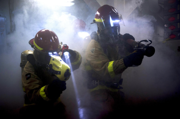 Zwei Feuerwehrleute in Schutzausrüstung sprühen Wasser auf eine rauchende Wand, mit einem Textbrett links und diversen Gegenständen rechts.