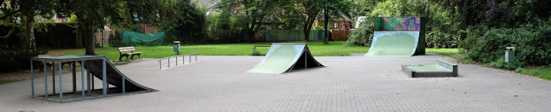 Ein Park mit einer zentralen Skaterrampe, umgeben von Bänken, Geländern, Grünflächen und Gebäuden im Hintergrund.