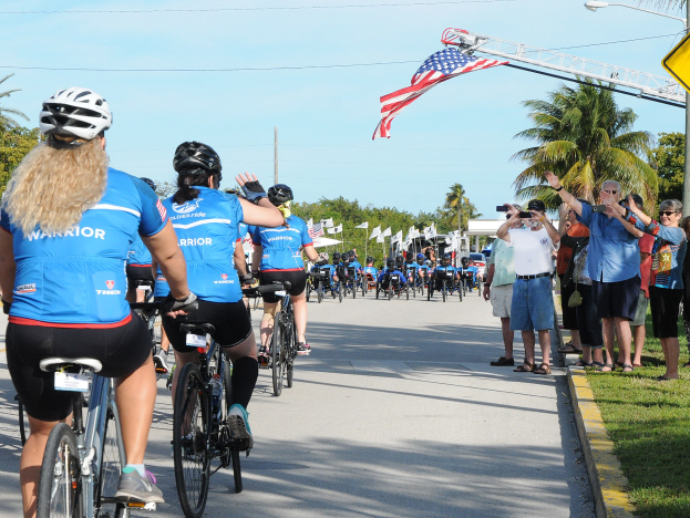 Radfahrer mit Helmen fahren auf einer Baumallee während eines Triathlon-Events, mit Zuschauern auf dem Rasen, die Kameras und Handys halten.