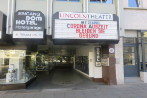 Das Lincoln Theater in Berlin, Deutschland, ein Gebäude mit Glasfenstern und -türen und einer Tafel mit Text darauf, das verschiedene Objekte im Inneren zeigt und den Eindruck einer pulsierenden Stadtlandschaft vermittelt.