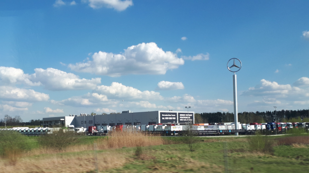 Ein Mercedes-Benz-Werk in Deutschland mit Fahrzeugen auf der Straße, Grünflächen, Gebäuden und einem bewölkten Himmel.