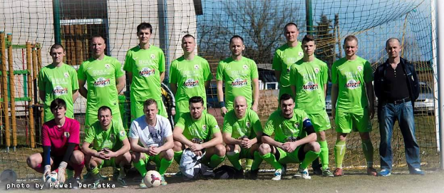 Eine Gruppe von Männern in grünen T-Shirts steht und hockt auf einem Fußballfeld mit einem Ball davor, ein Torpfosten rechts und ein Hintergrund aus Bäumen, Gebäuden, Pfählen, Fahrzeugen und Himmel.
