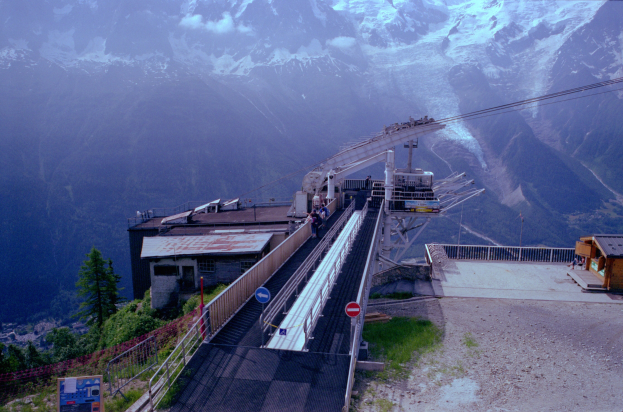 Seilbahn fährt einen schneebedeckten Berg mit Straße, Schildern, Geländern, Vegetation, Häusern und Menschen am Fuße hoch.