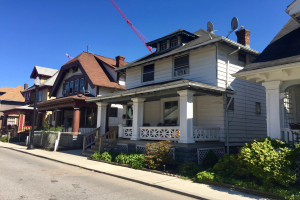 Eine Reihe von Häusern mit architektonischen Details wie Fenstern, Säulen, Geländern, Treppen, Dächern, mit einem Straßenpfahl, Pflanzen, einem Zaun, einem Weg, Bäumen und einem bewölktem Himmel, mit einem Baukran auf einem Dach, der laufende Bauarbeiten anzeigt.