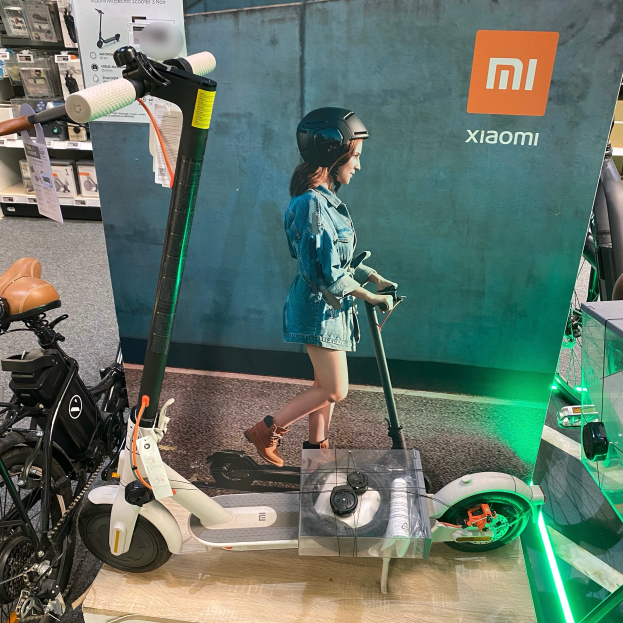 Eine Frau mit Helm fährt ein Xiaomi M365-Elektroroller in einem Geschäft, rechts daneben eine Glasvitrine mit verschiedenen Gegenständen und im Hintergrund Schautafeln mit Text und Bildern.