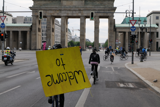 Eine Gruppe von Radfahrern fährt am Brandenburger Tor in Berlin vorbei, einer hält ein gelbes Schild, mit Laternenpfählen, Verkehrsampeln, Gebäuden und Bäumen im Hintergrund und einem klaren blauen Himmel.