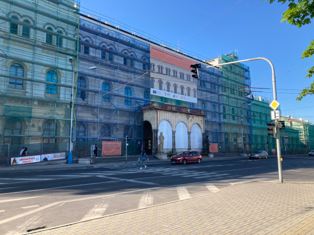 Ein großes Gebäude mit zahlreichen Fenstern, umgeben von Fahrzeugen, Fußgängern und verschiedenen Straßenelementen, mit Gerüsten, die anlaufende Renovierungsarbeiten unter einem klaren blauen Himmel anzeigen.